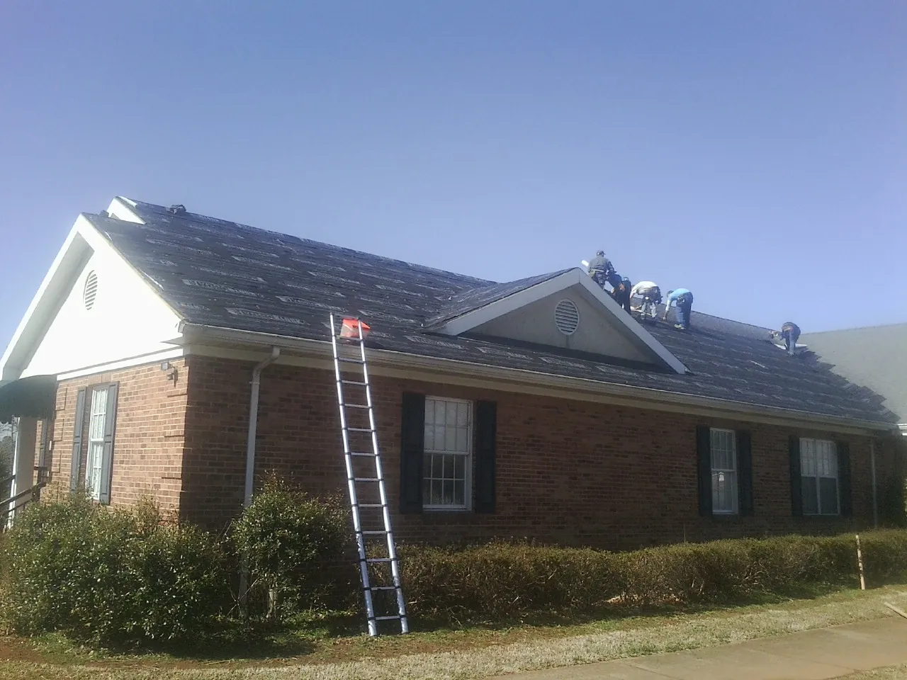 Crew tearing shingles off a brick ranch roof with a ladder set — Emergency Roof Repair work by our crew in East Nassau