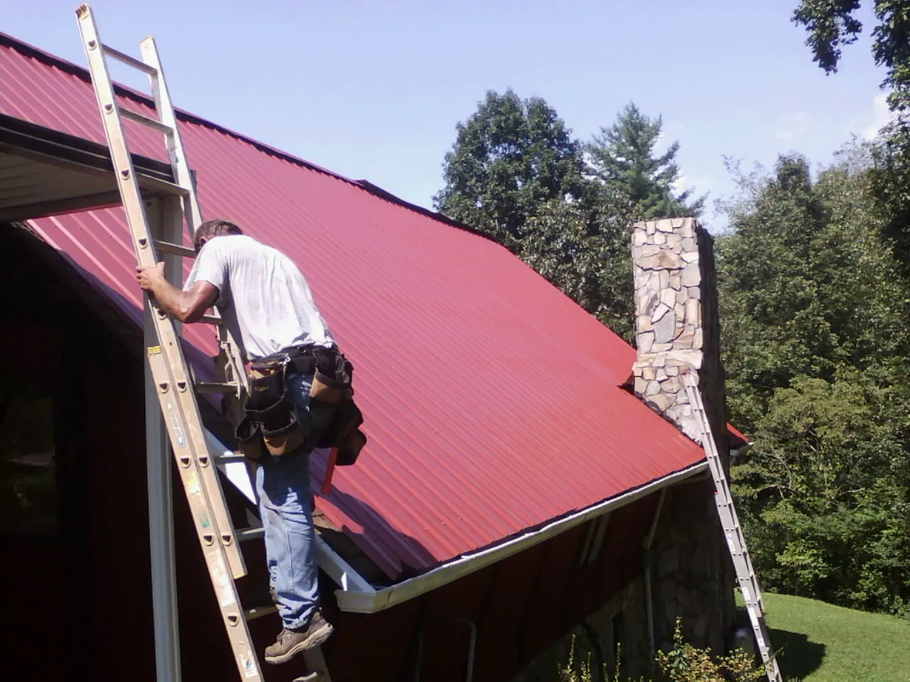 Roofer on a ladder beside a red metal roof and stone chimney — Soffit & Fascia Repair by Roofing Brothers in East Nassau